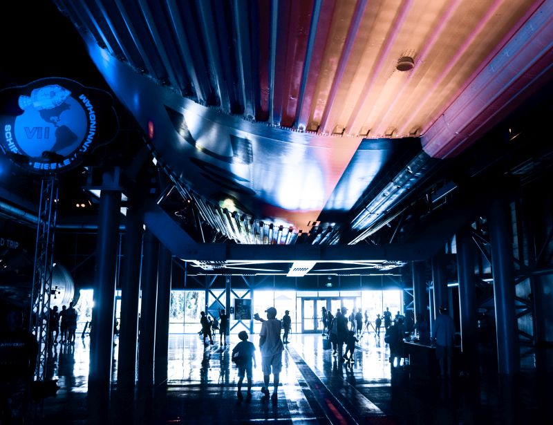 Kai and Kenny return toward the entranceway to the Great Hall, beneath the first stage of the Saturn V, headed toward the Apollo Command/Service module, which can be seen partially, at floor-level, beneath the Apollo VII mission patch above, on the far left of this frame.